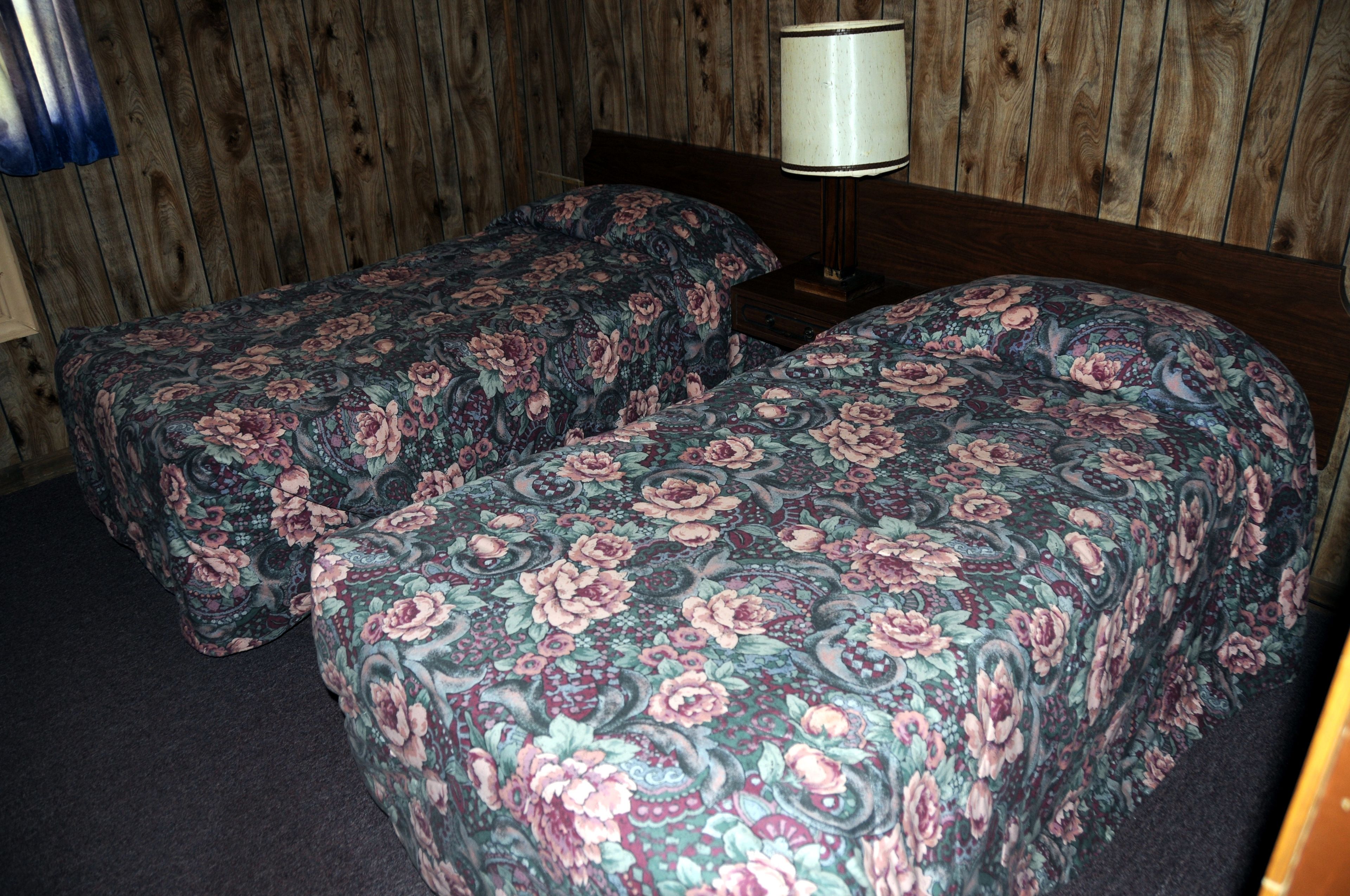 A room with two single beds with floral bedding, a wooden headboard, and a bedside lamp.