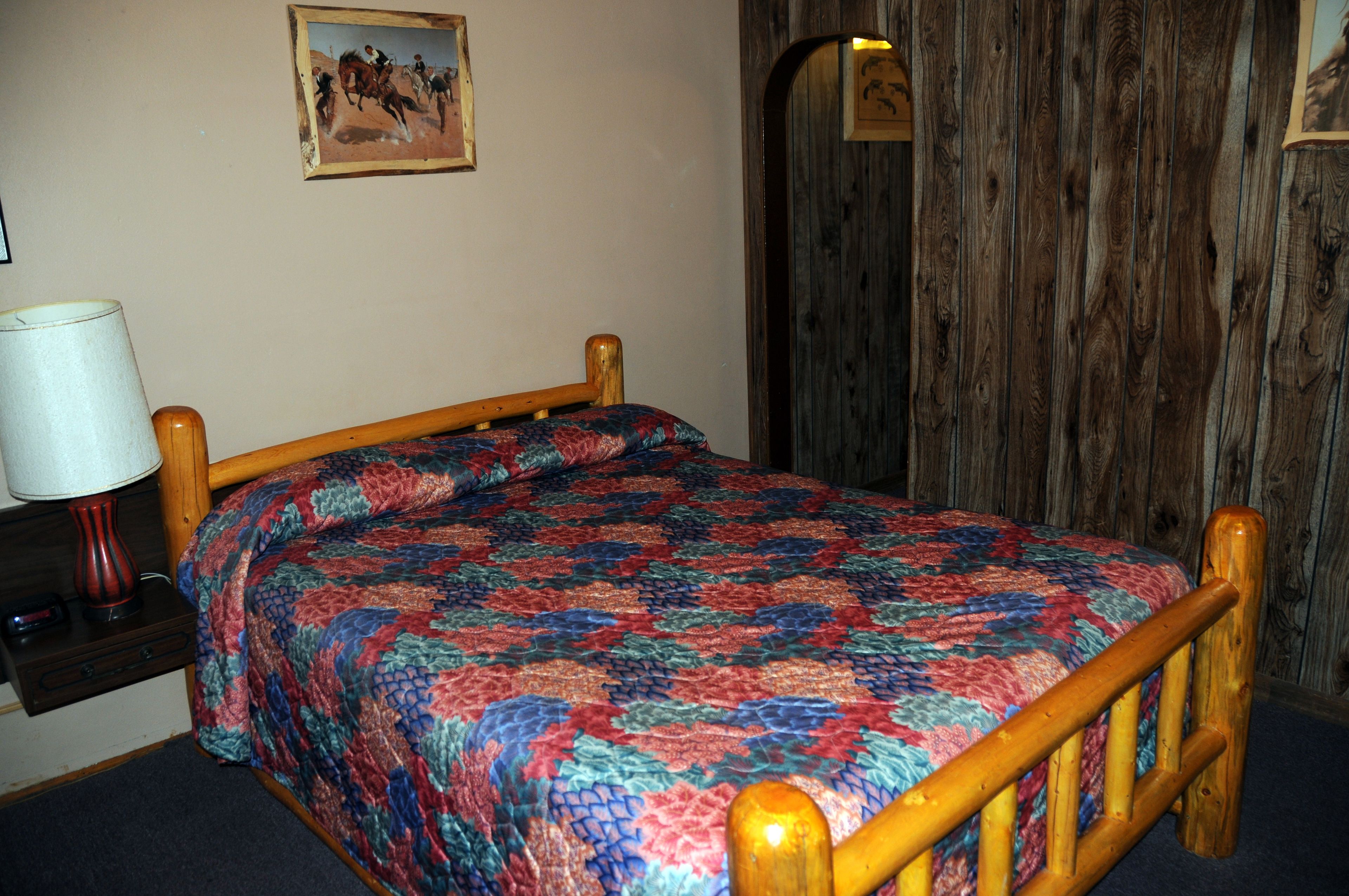 Una cama con un edredón colorido, un cabecero de madera, una mesita de noche con una lámpara y un cuadro en la pared.