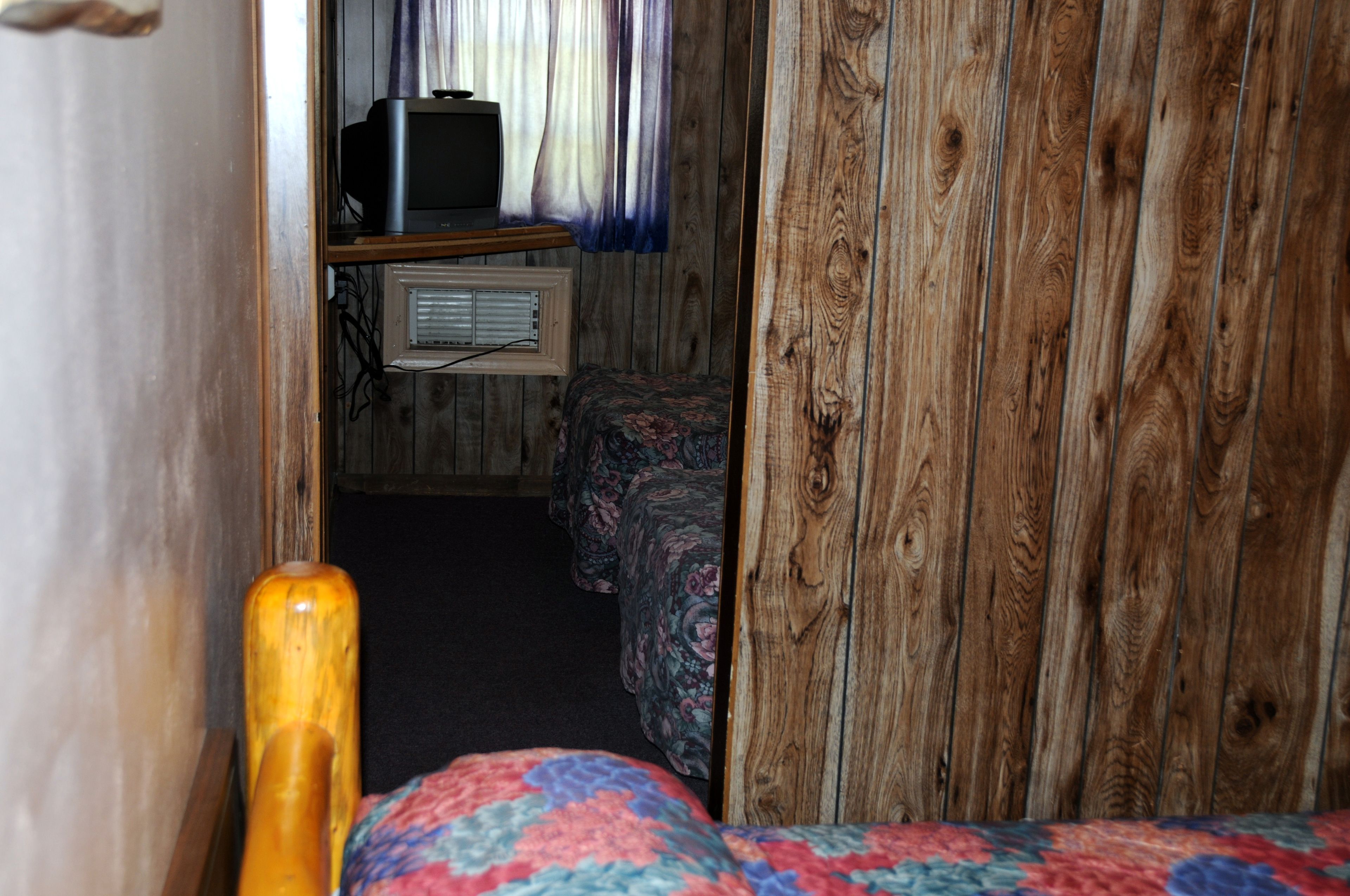 Una habitación pequeña con pared de madera, una cama con colcha estampada, una mesita con televisor y un radiador.