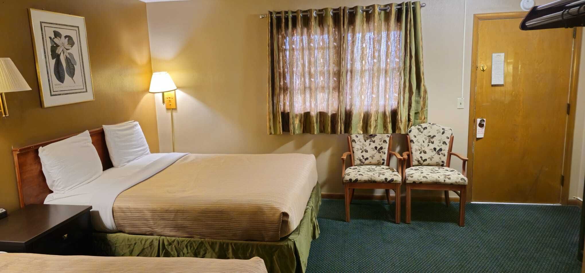 Ein Hotelzimmer mit einem Bett, zwei Stühlen, einer Lampe, einem gerahmten Bild und einer Tür.
