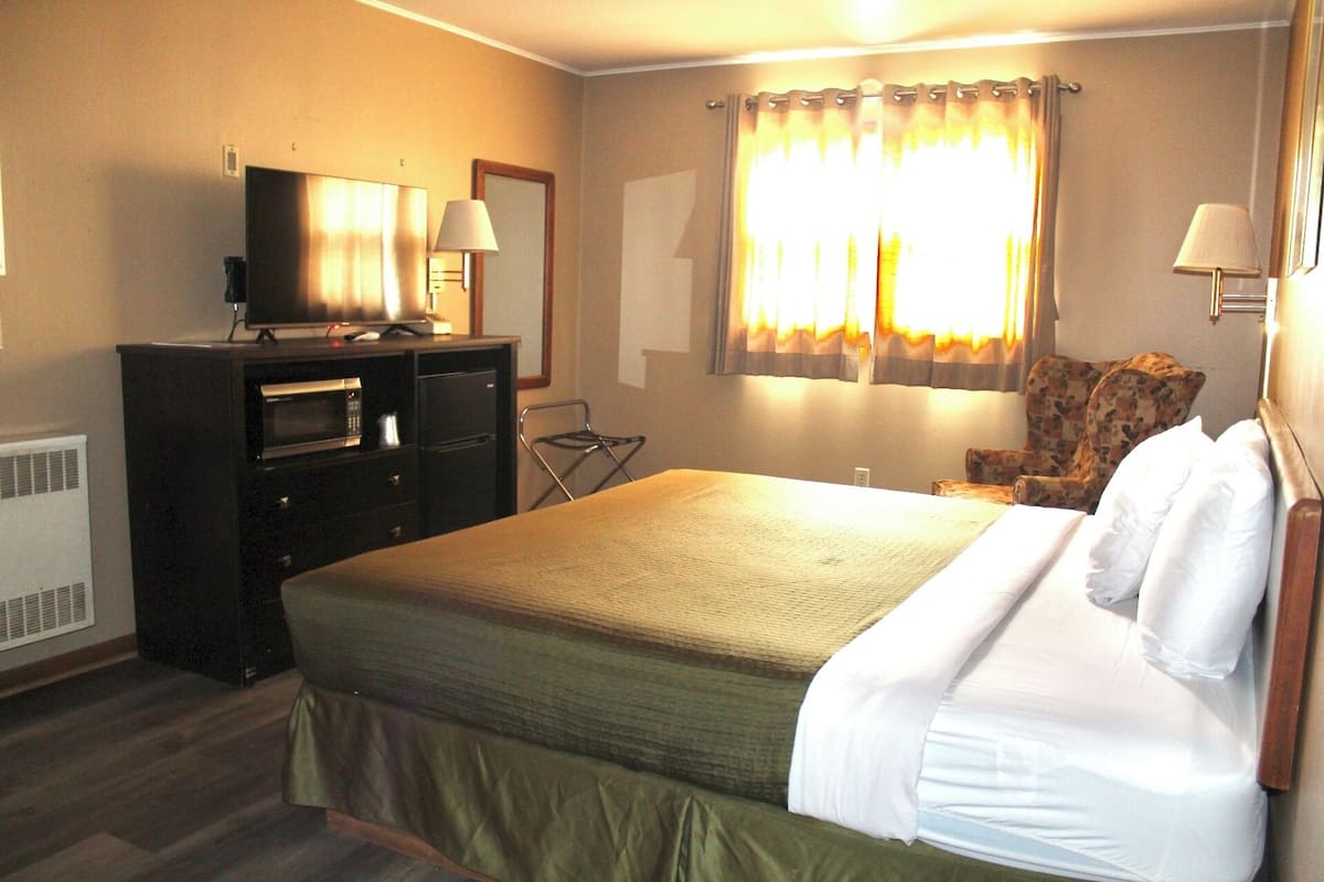 Ein Hotelzimmer mit einem Bett, einem Schreibtisch mit Computer, einem Stuhl, einem Fenster mit Vorhängen und einem an der Wand befestigten Fernseher.