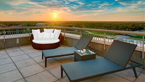 Un balcón con zona de estar, que incluye un sofá y dos sillones, con vistas a un paisaje urbano al atardecer.