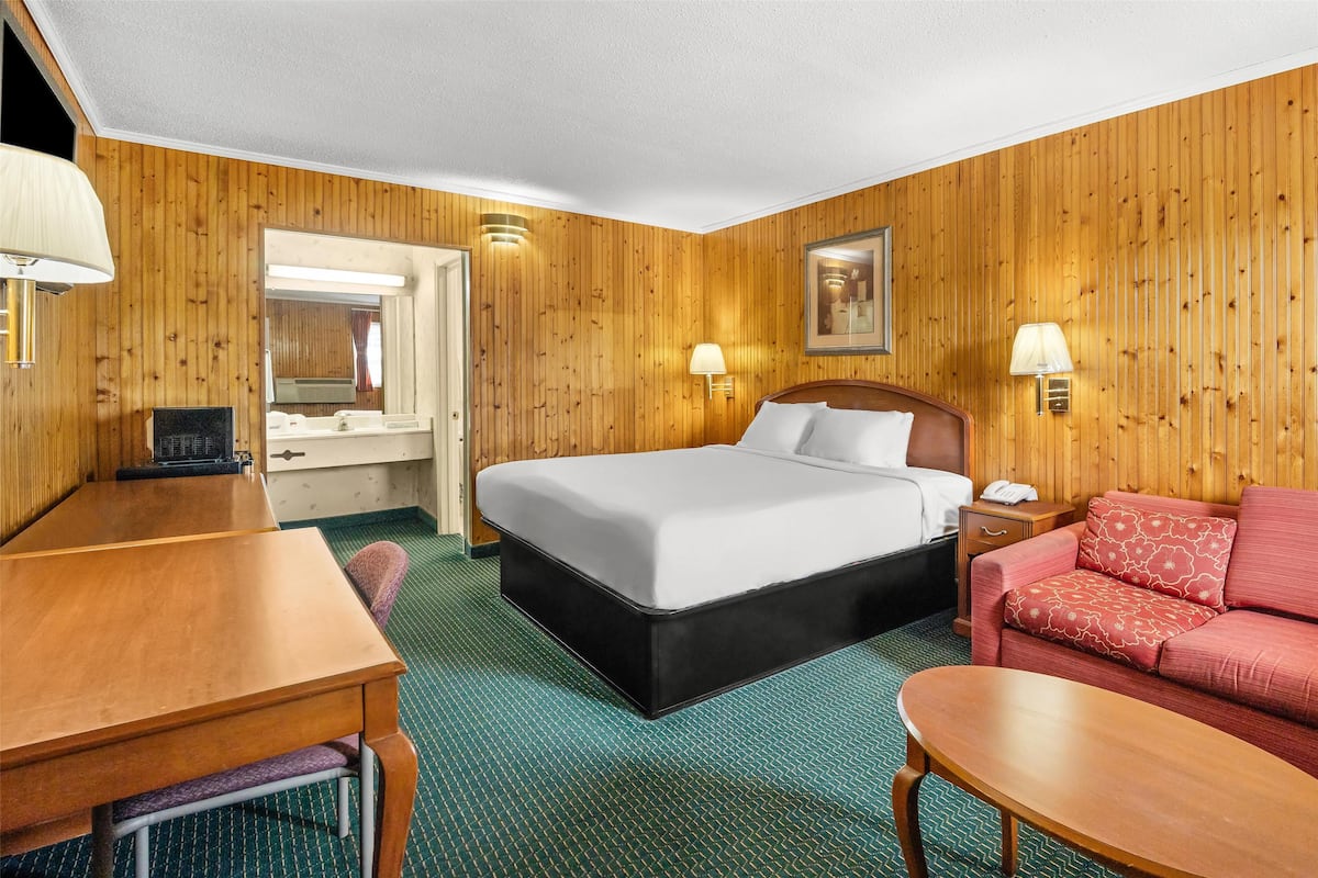Una habitación de hotel con pared de madera, una cama con ropa blanca, un sofá rojo, un escritorio de madera y una alfombra verde.