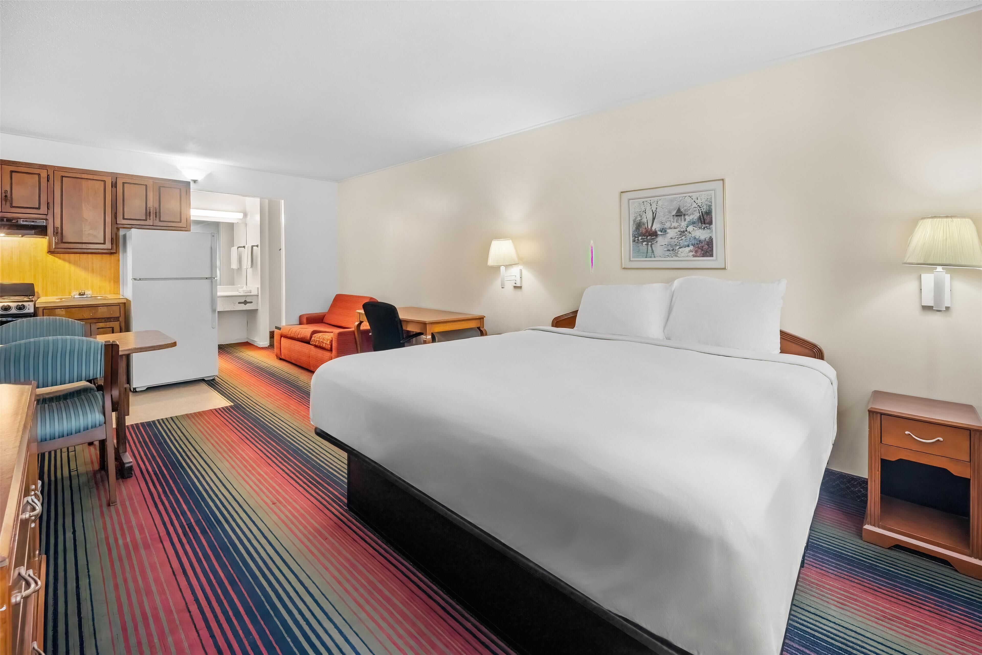 Habitación de hotel con cama, escritorio con silla, una kitchenette pequeña y una alfombra a rayas colorida.
