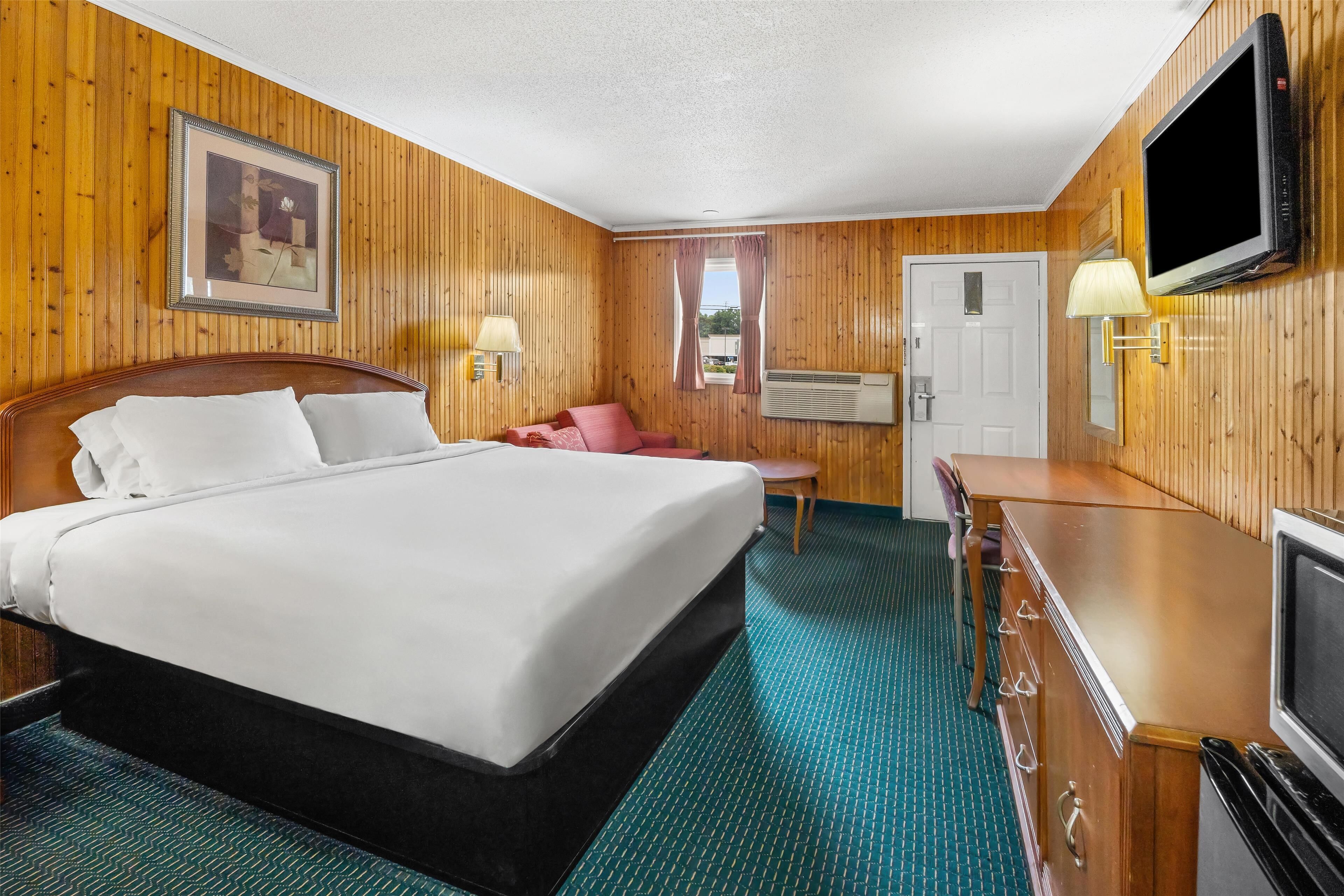 A hotel room with a wooden paneled wall, a bed with white linens, a television, a desk, and a chair.