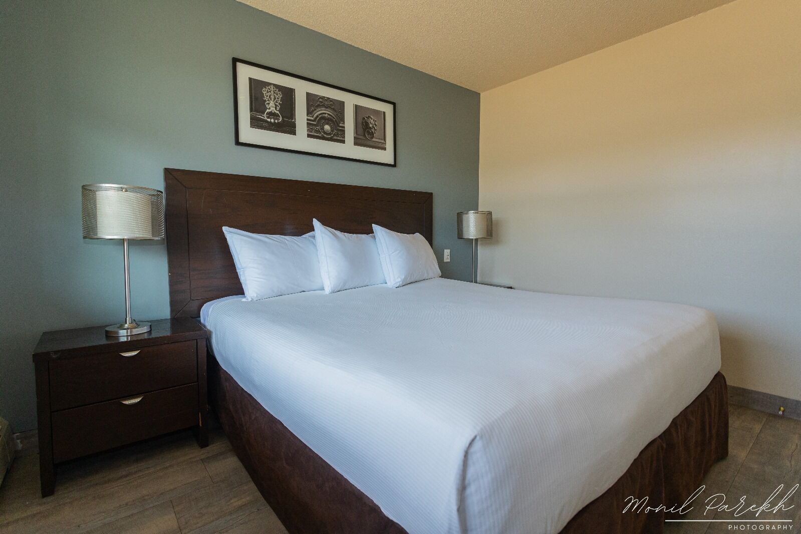 A neatly made bed with white linens, a wooden headboard, and a nightstand with a lamp. There is a framed picture above the bed.