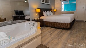 A hotel room with a large bathtub, a bed with a brown headboard, two bedside lamps, and a window with a view of the sky. - Empire Inn & Suites (Red Deer)