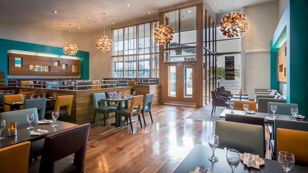 Un interior de restaurante moderno con suelo de madera, varias zonas de estar y ventanas amplias que dejan entrar luz natural.