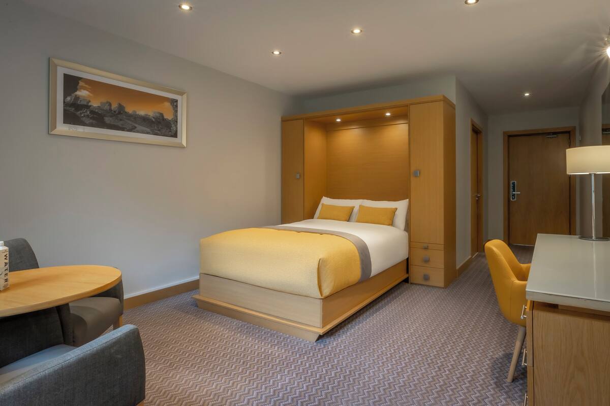 A hotel room with a bed, a desk, a chair, a mirror, and a painting on the wall.