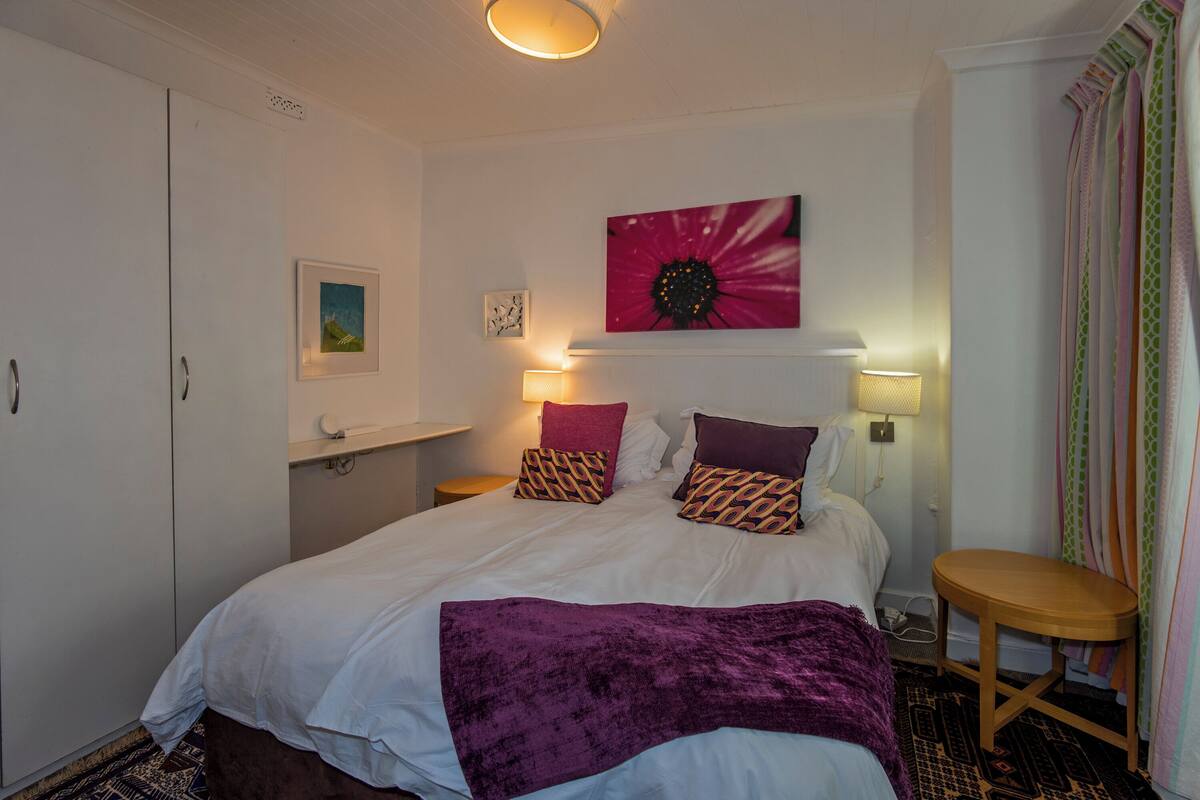 A bedroom with a bed, two bedside lamps, a nightstand, a chair, a window with curtains, and artwork on the wall.