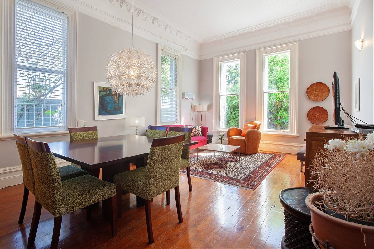 A dining area with a wooden table and chairs, a chandelier, and a view of greenery outside.