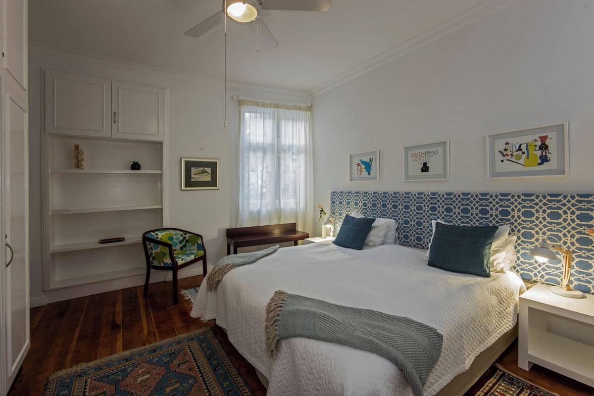 A bedroom with a large bed, a chair, a nightstand, a desk, and a window with curtains.