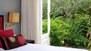 Une chambre d’hôtel avec un lit, un oreiller rouge, un rideau blanc et une vue sur un jardin où poussent différentes plantes et un arbre.