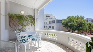 Un balcon avec des meubles en osier blancs, un mur blanc et une vue sur des immeubles et des arbres.