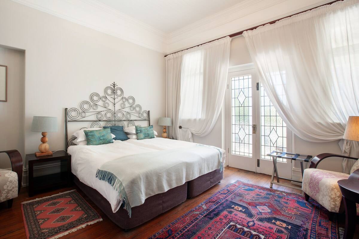 A bedroom with a large bed, a nightstand with a lamp, a chair, a small table, and a patterned rug.