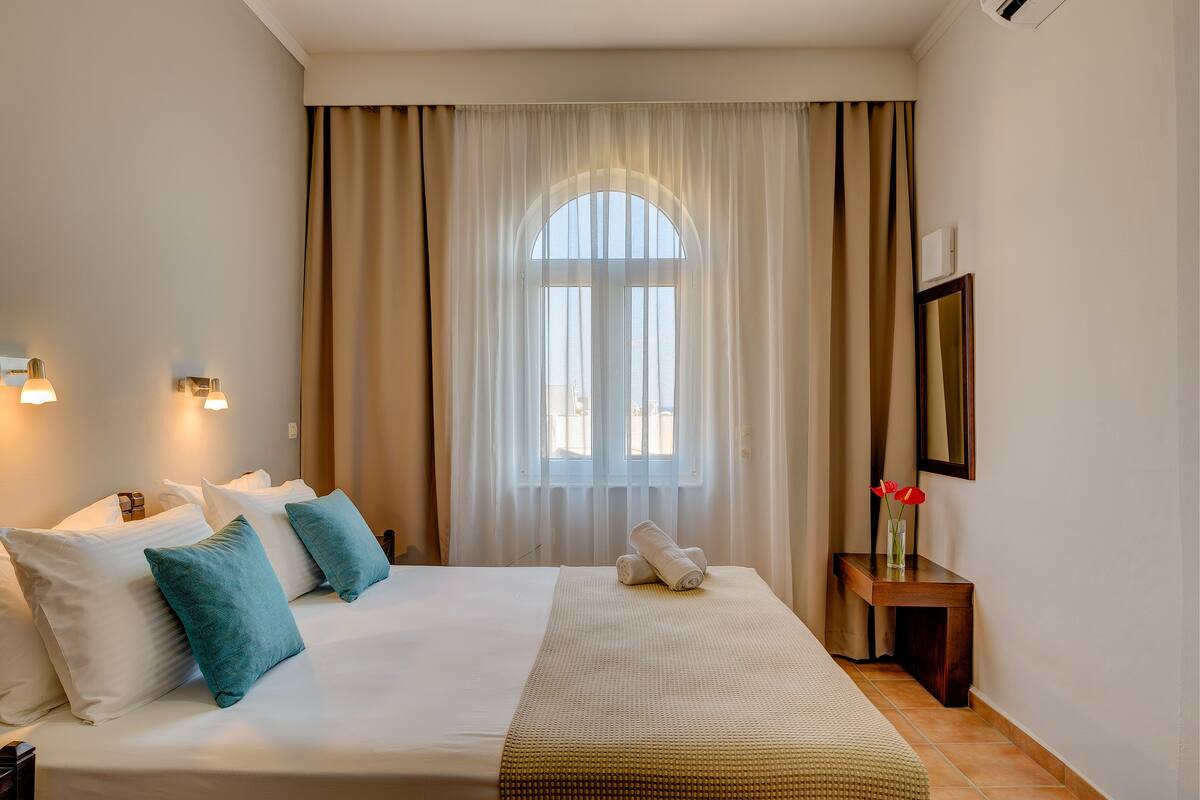 A hotel room with a bed, pillows, a bedside table with a vase of flowers, and a window with curtains.