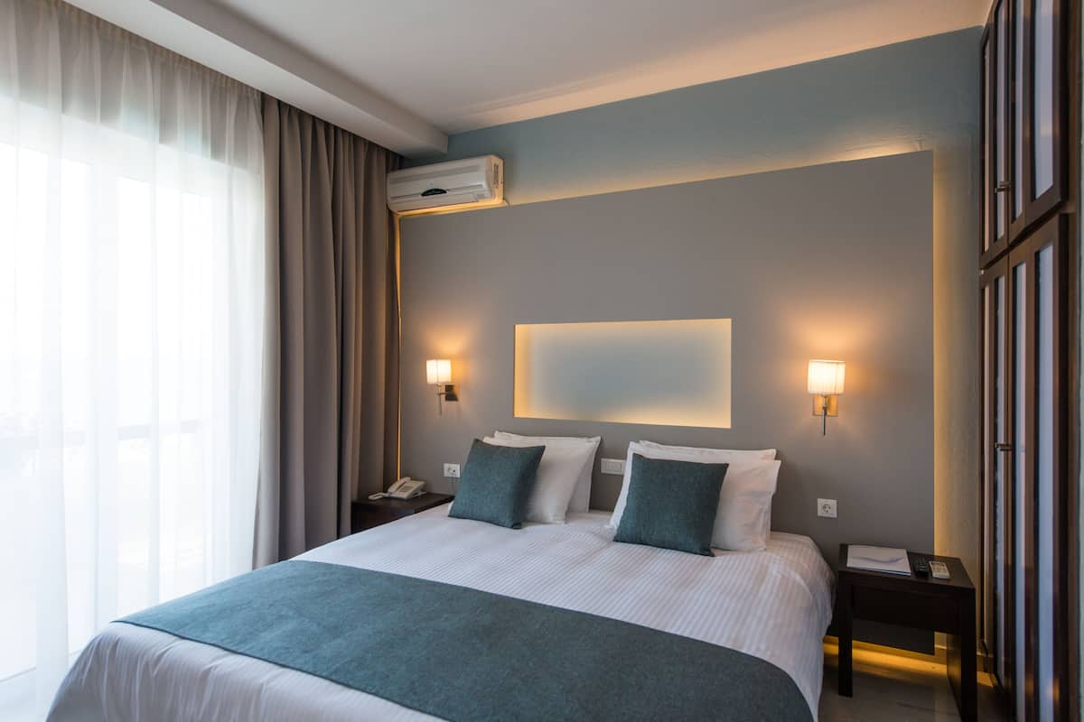 A hotel room with a large bed, two bedside tables, a nightstand, a wall-mounted air conditioner, and a window with curtains.