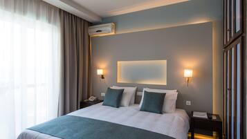 A hotel room with a large bed, two bedside tables, a nightstand, a wall-mounted air conditioner, and a window with curtains.