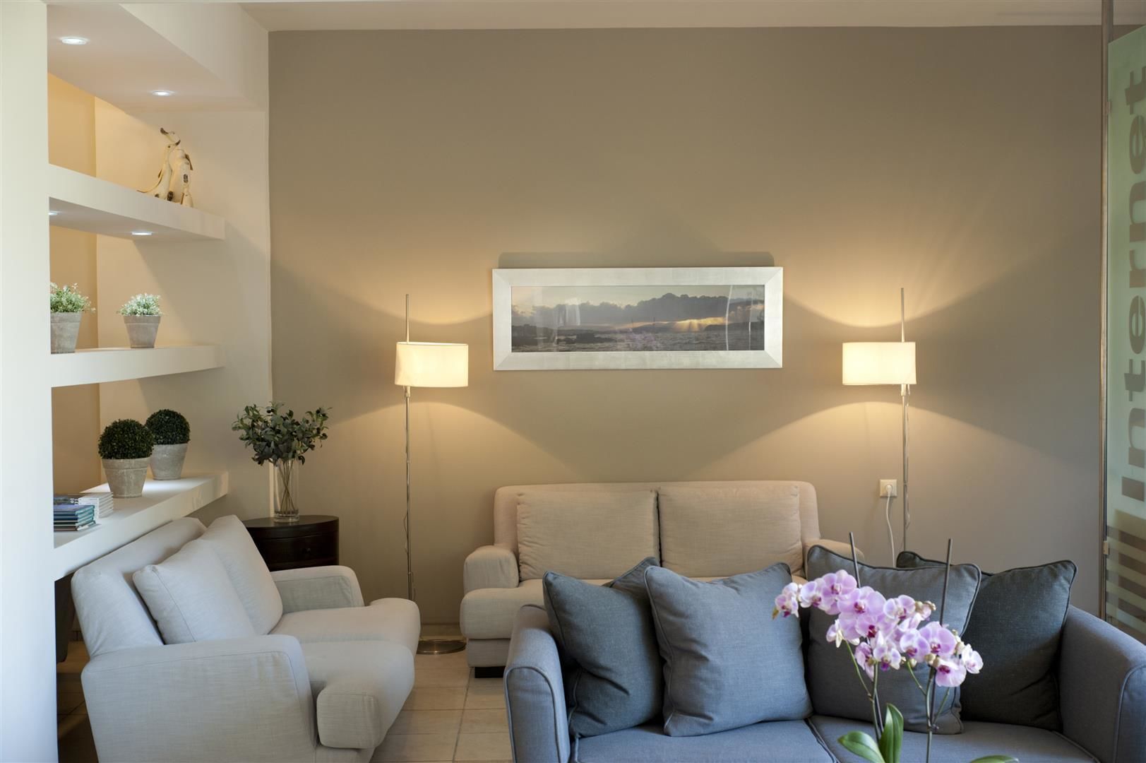 A living room with a beige sofa, a grey sofa, and a floor lamp. There is a framed picture on the wall and a small table with a vase of flowers.