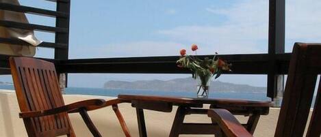 A balcony with two wooden chairs and a small table, overlooking a scenic view of the sea and distant mountains.