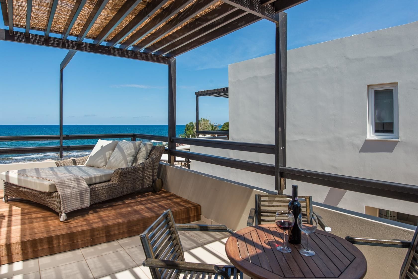 A balcony with a wicker sofa, a wooden table with wine glasses, and a view of the ocean.