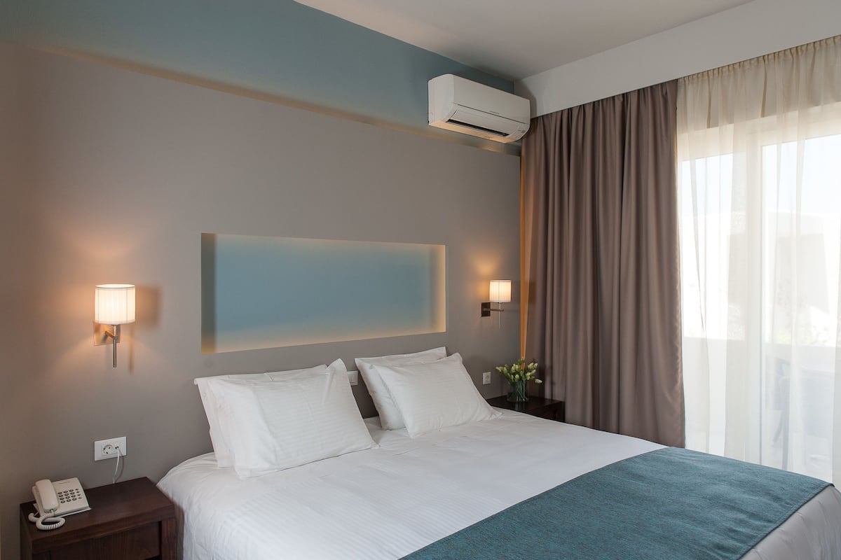 A hotel room with a bed, a nightstand with a phone, an air conditioner, and a painting on the wall.