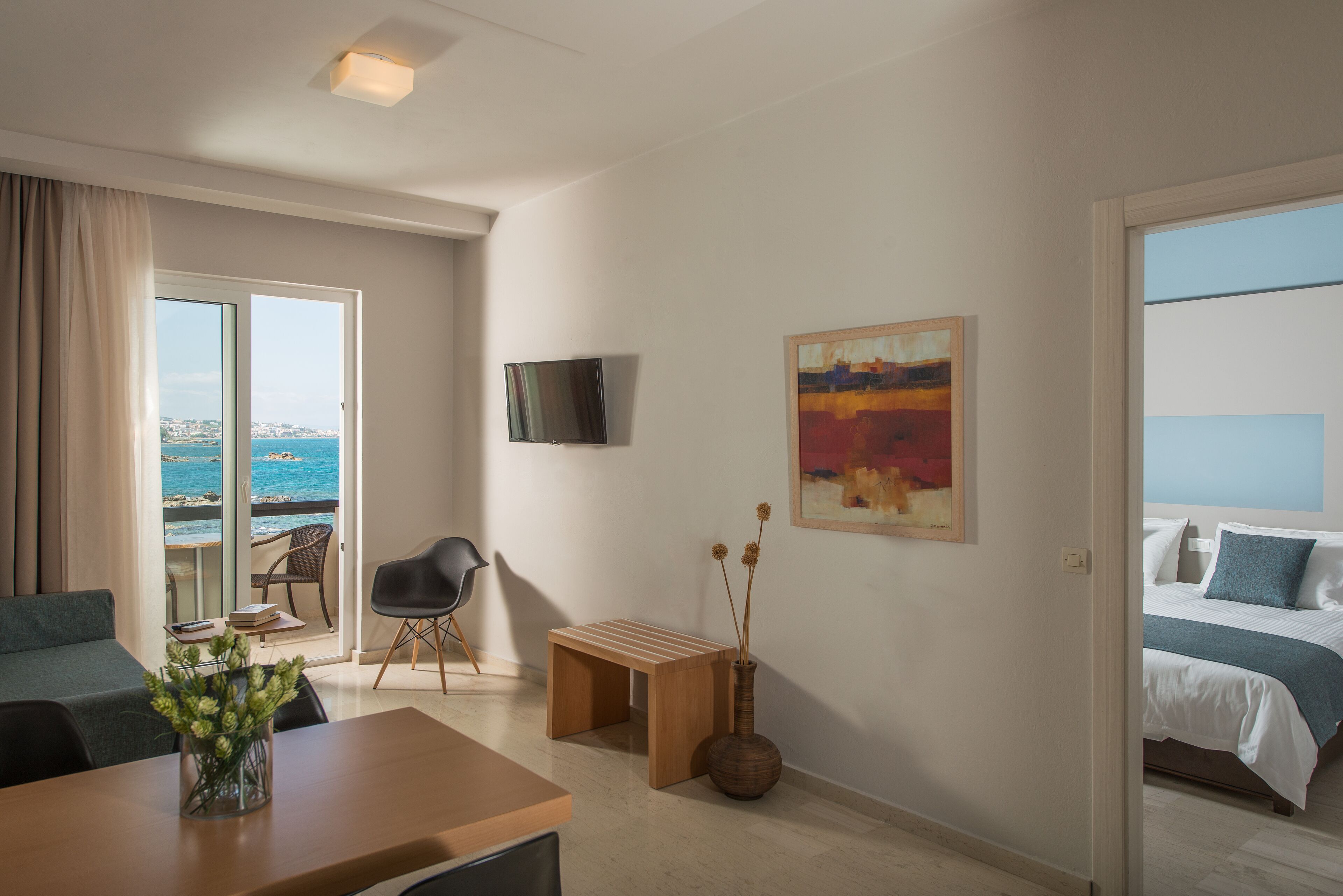 A hotel room with a balcony, a TV, a painting, a bed, and a dining area with a table and chairs.