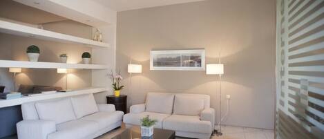 Una sala de estar moderna con dos sofás blancos, una mesa de centro de vidrio y estanterías con objetos decorativos.