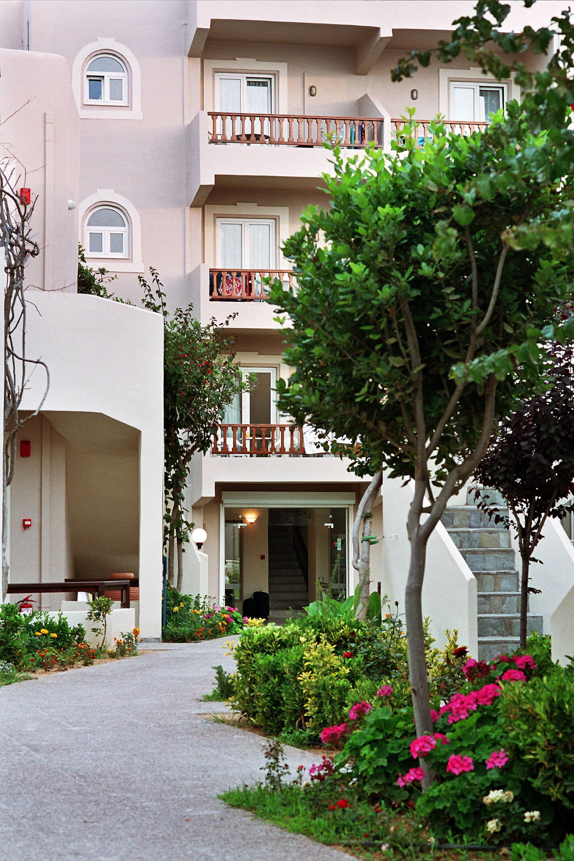 Un immeuble à plusieurs étages doté de balcons, un jardin avec différentes plantes et fleurs, et un chemin menant à l’entrée.