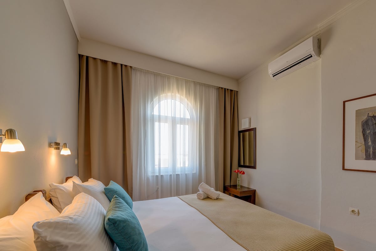 A hotel room with a large bed, a window with curtains, an air conditioning unit, a bedside table with a lamp, and a framed artwork on the wall.