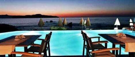 Una piscina infinita con vistas al atardecer, mobiliario exterior con sombrillas y una terraza de madera.