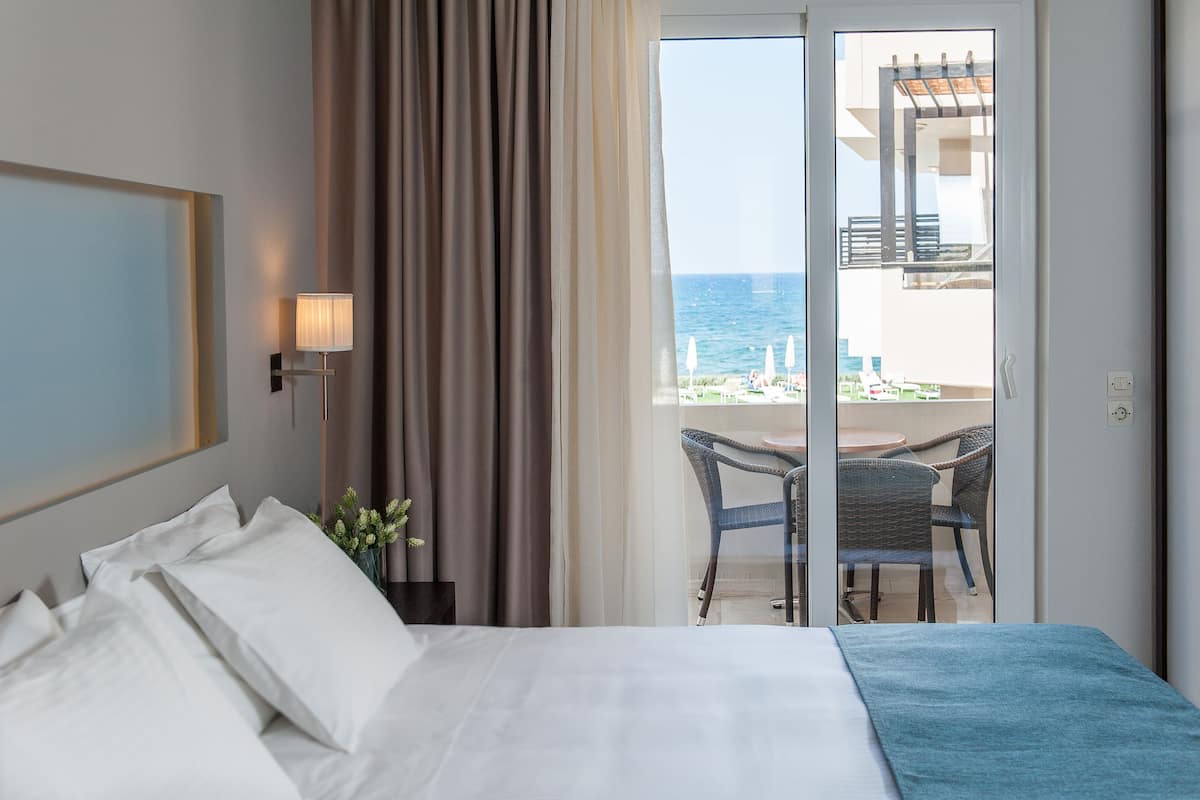 A hotel room with a bed, a nightstand, a lamp, a window with curtains, and a view of the sea and outdoor seating.