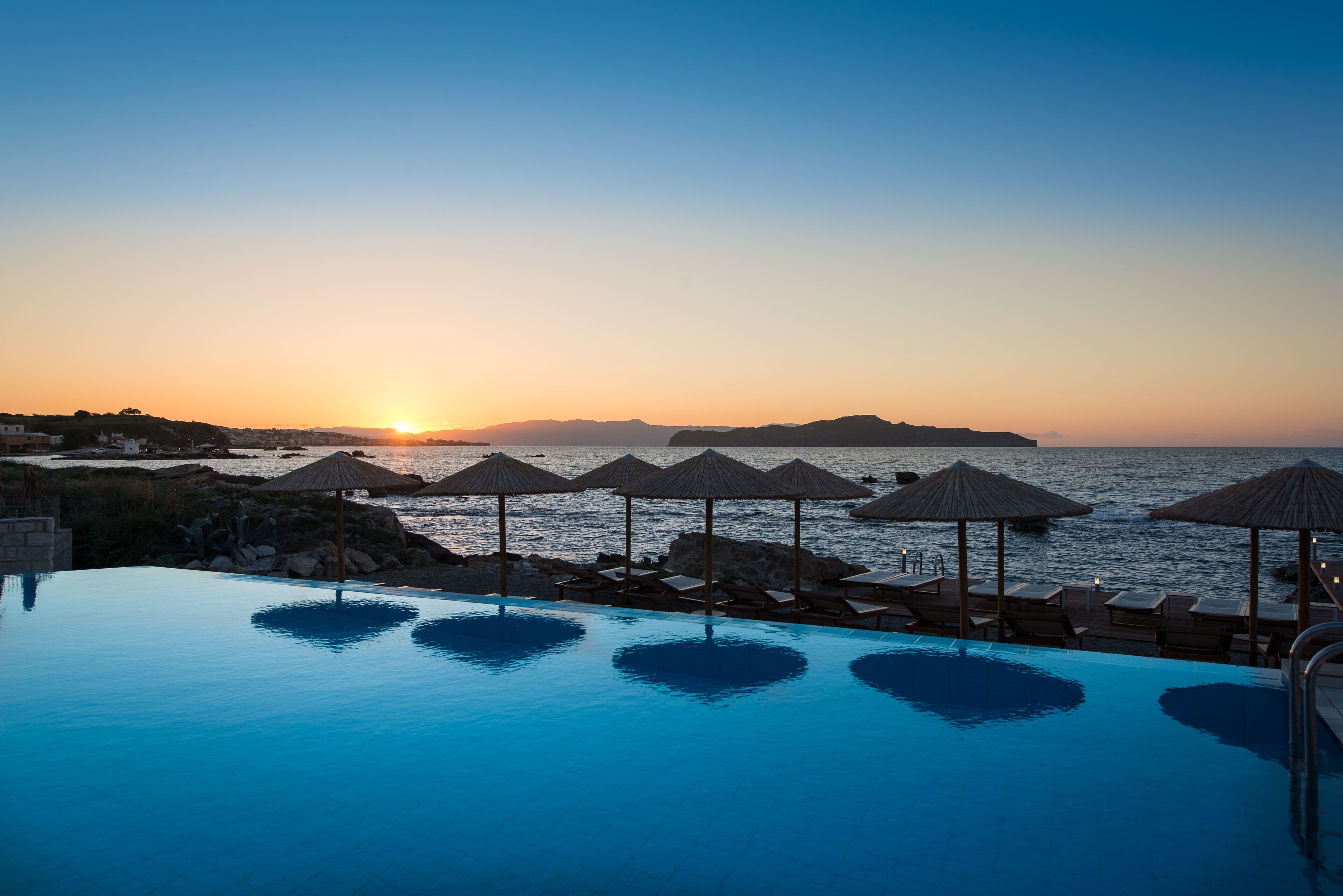 Ein Poolbereich mit Sonnenschirmen und Liegestühlen mit Blick auf das Meer bei Sonnenuntergang.