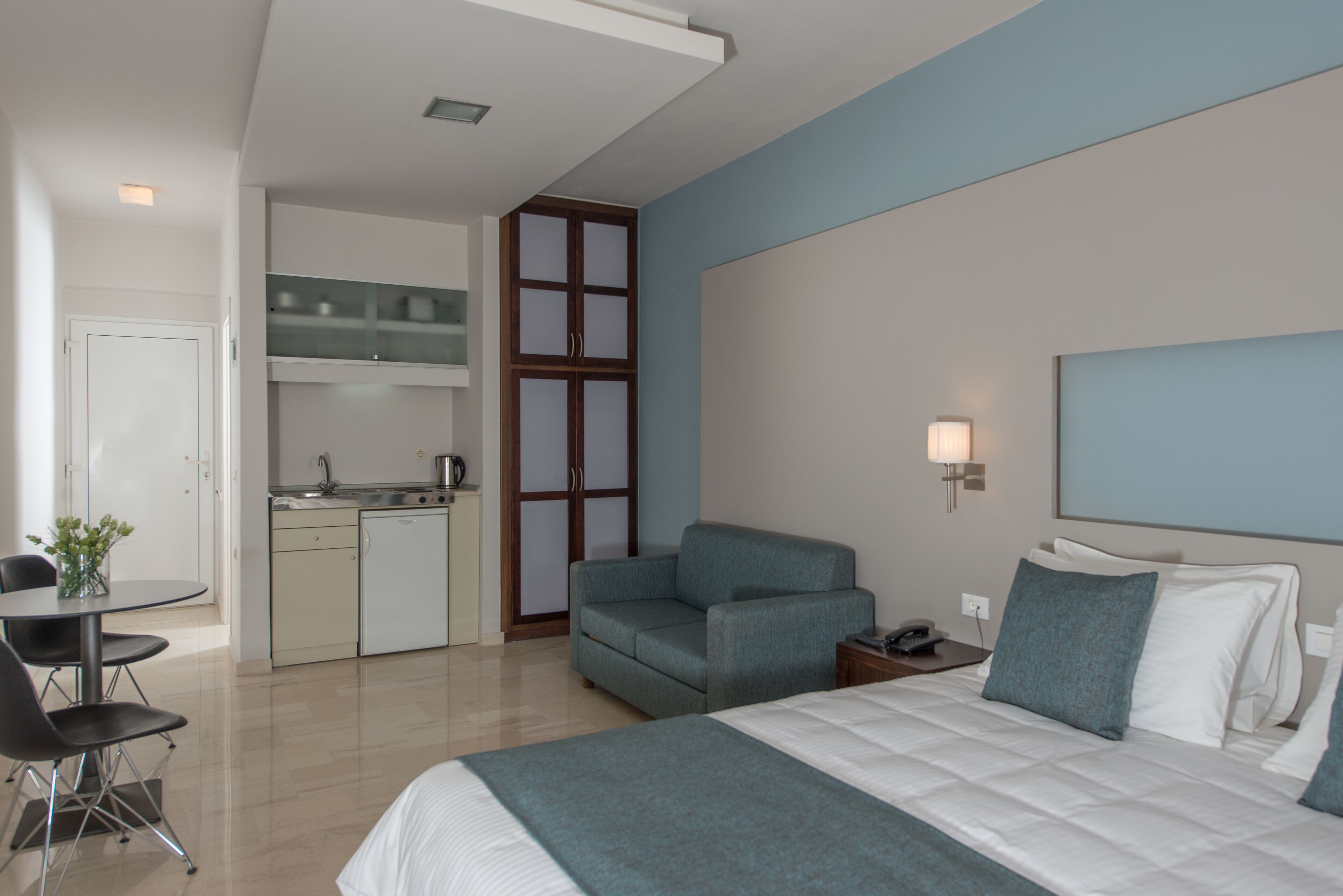 A modern hotel room with a bed, a sofa, a dining table, and a kitchenette.