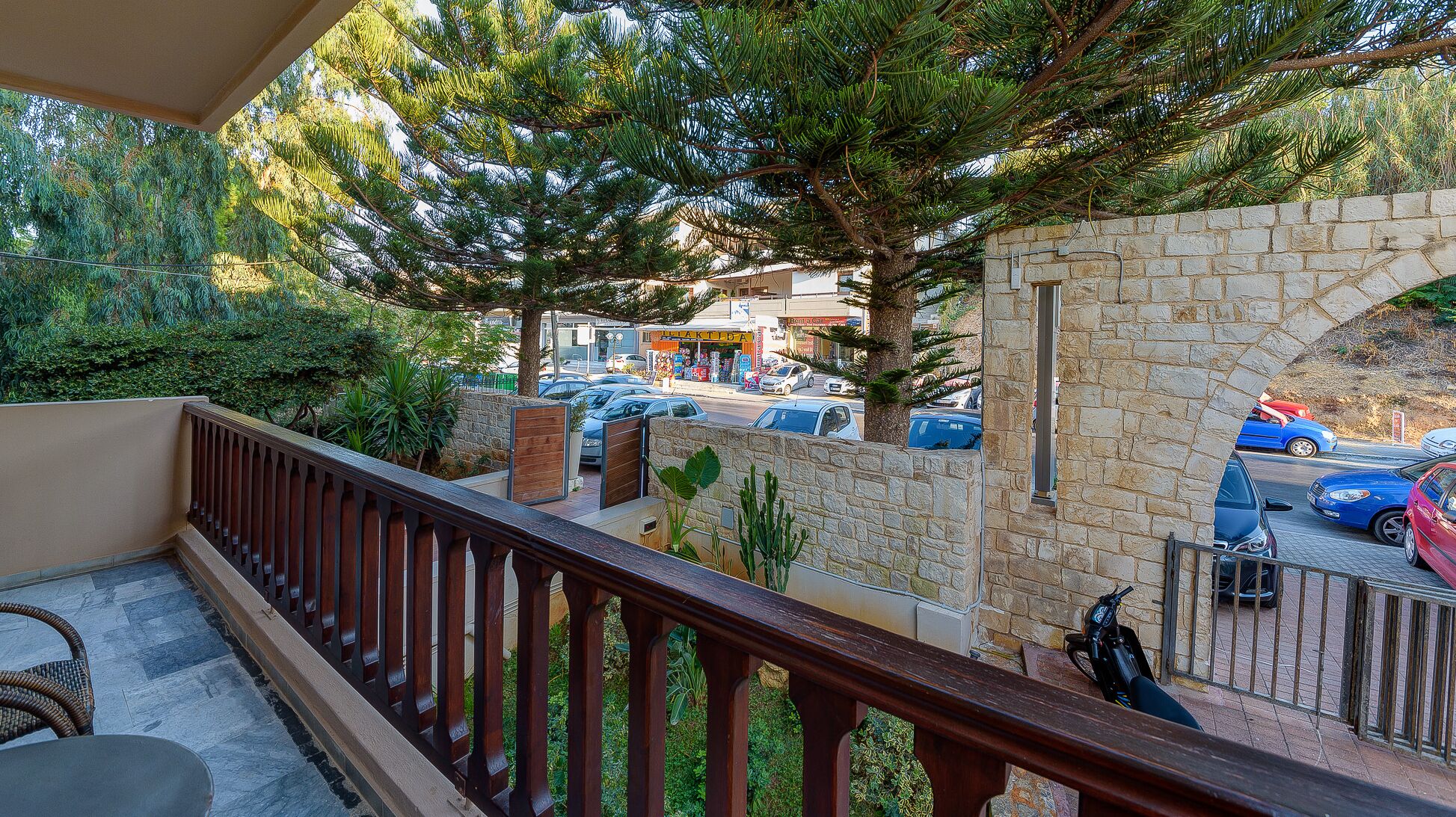 Un balcon avec un mur en pierre, une vue sur des voitures garées et un immeuble avec des boutiques en arrière-plan.