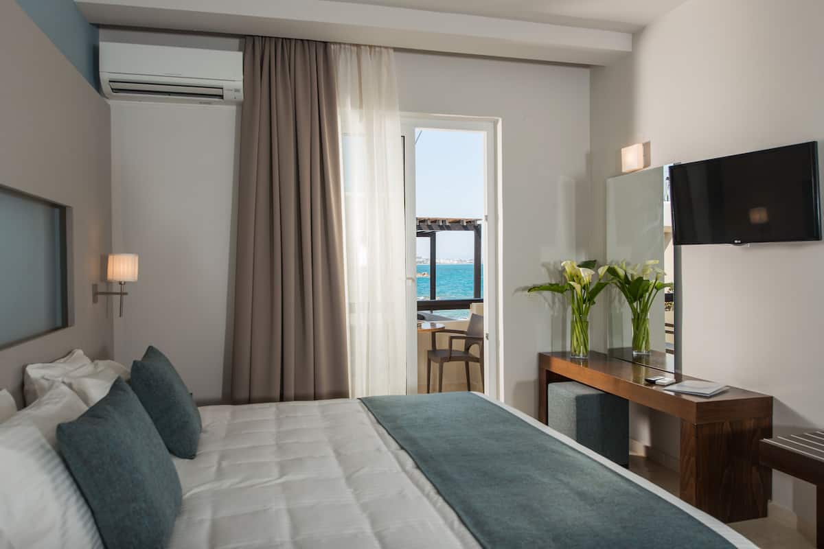 A hotel room with a bed, a desk, a TV, and a view of the sea through an open door.