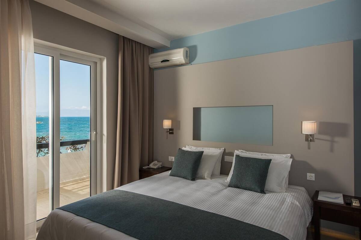 A hotel room with a large bed, two bedside tables, a nightstand, a wall-mounted air conditioner, and a view of the ocean through a sliding door.