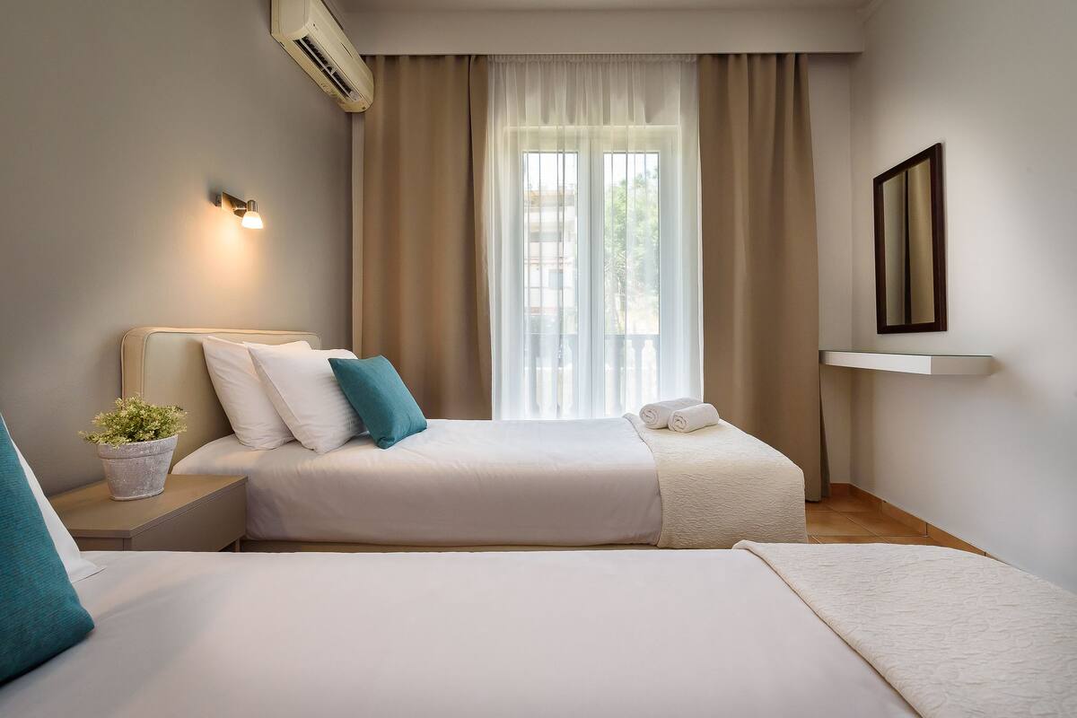 A hotel room with two beds, a potted plant, a wall-mounted air conditioner, and a window with curtains.
