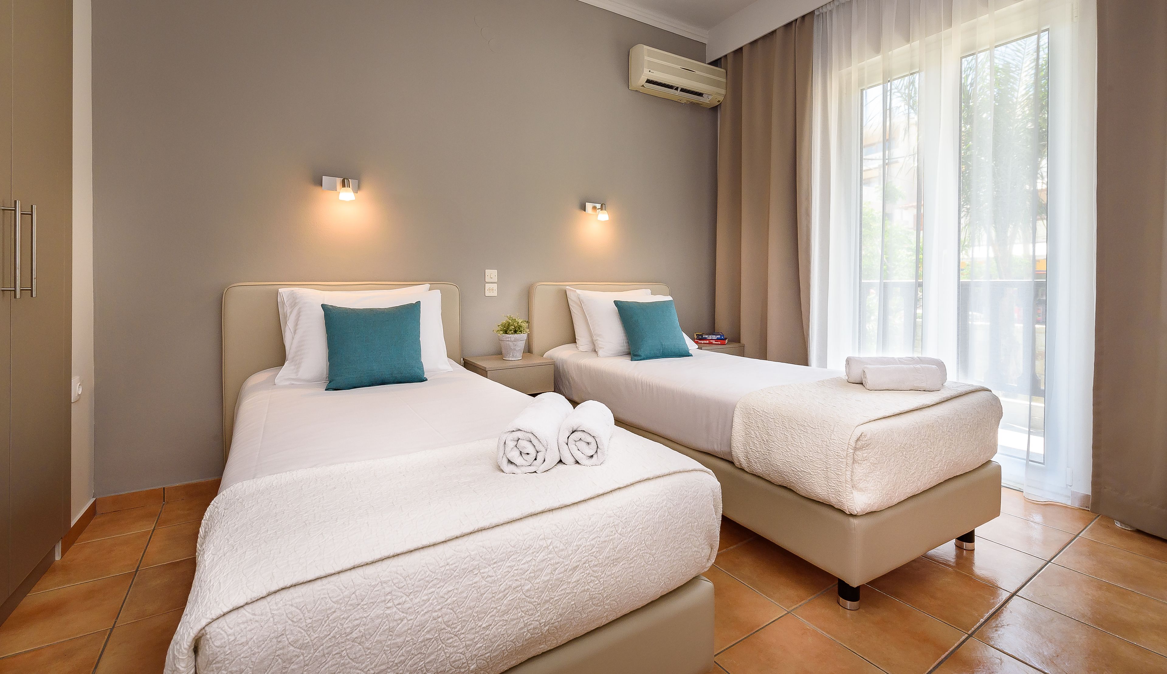 Habitación de hotel con dos camas, cada una con ropa blanca y almohadas color turquesa, una mesita de noche con una planta, y un ventanal con cortinas traslúcidas.