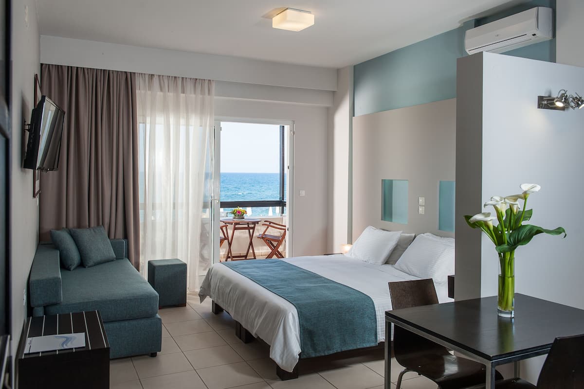 A hotel room with a bed, a sofa, a dining table, and a view of the sea.