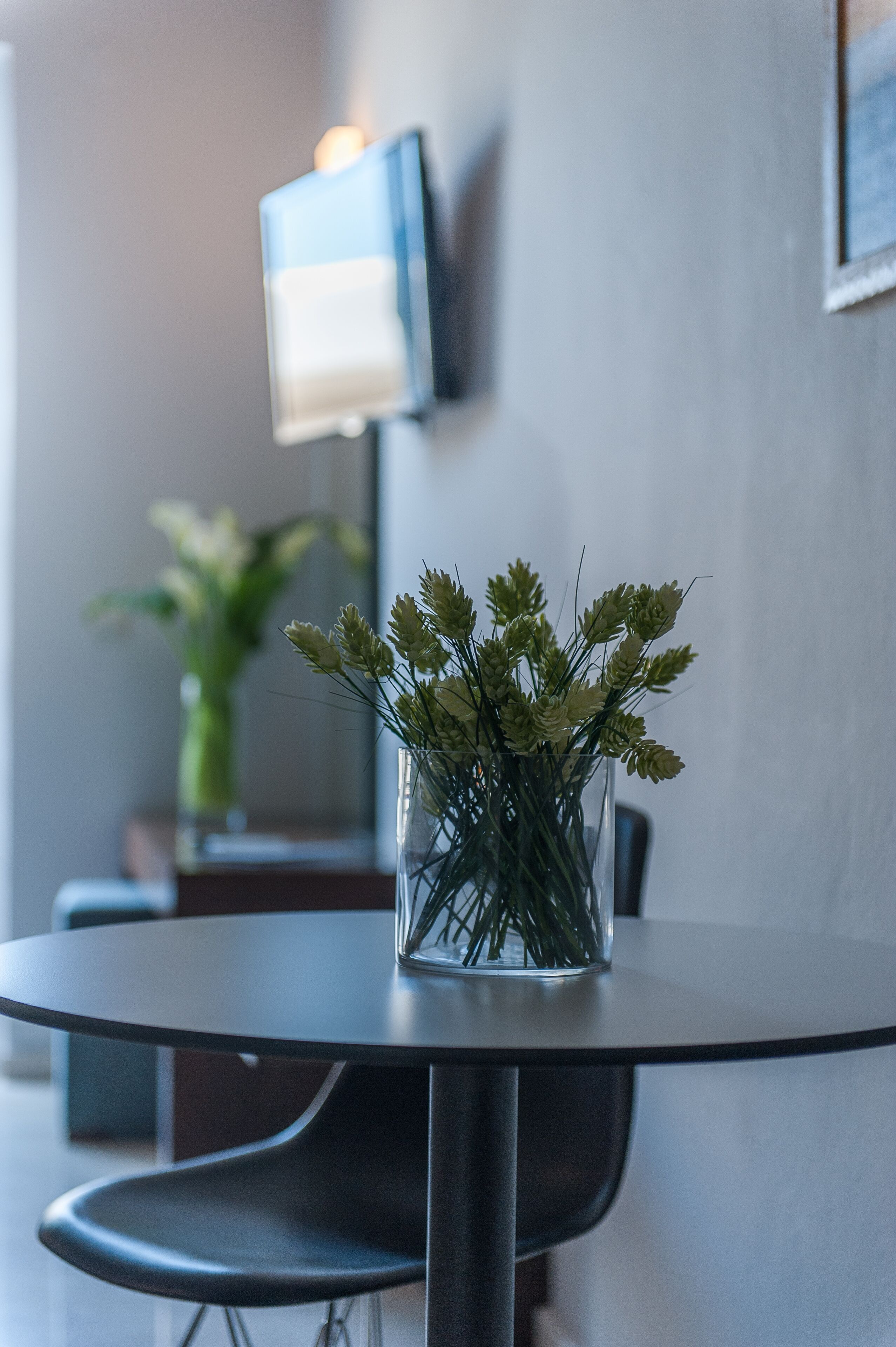 A round table with a clear glass vase containing green plants, a modern lamp, and a framed picture on the wall.
