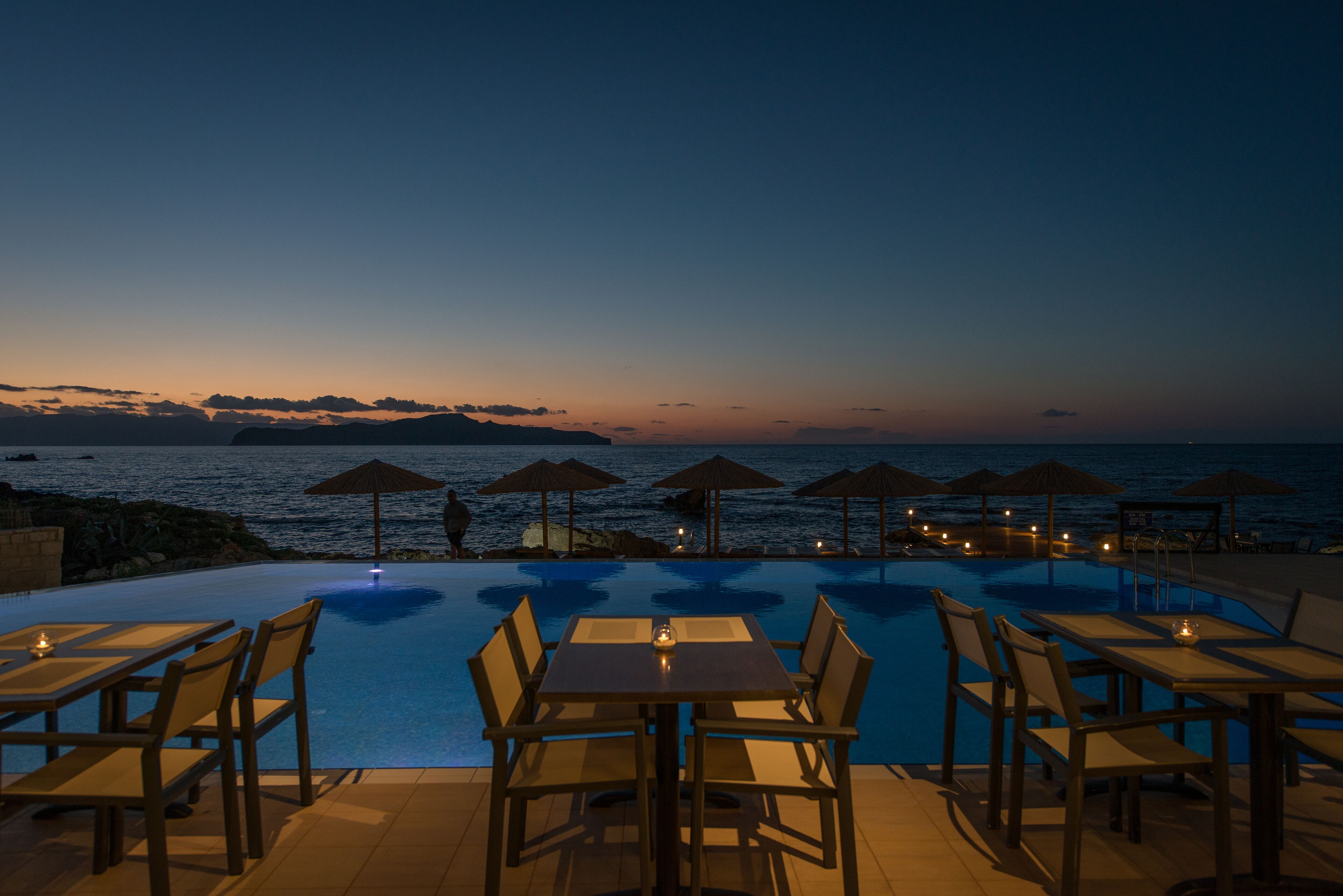 A poolside area with tables and chairs, umbrellas, and a view of the sea at dusk.