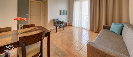 A living room with a sofa, a dining table with chairs, a TV mounted on the wall, and a door leading to another room.