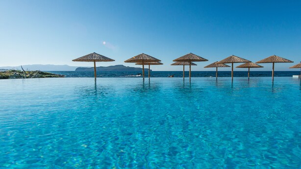 A clear blue infinity pool with thatched umbrellas and a view of the sea and mountains in the background. - Corinna Mare (Chania)