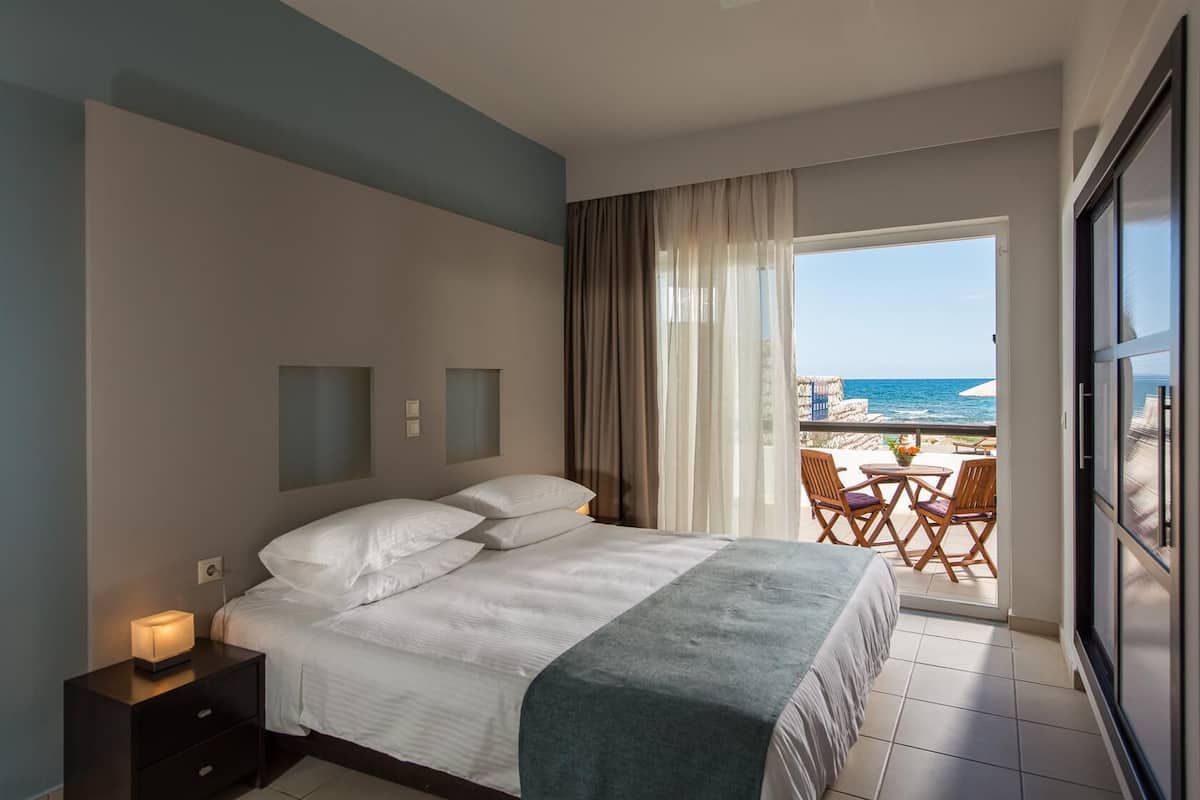 A hotel room with a bed, a nightstand, a window with a view of the beach, and a balcony with chairs.