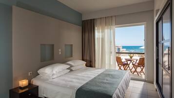 Ein Hotelzimmer mit Bett, Nachttisch, Fenster mit Meerblick und Balkon mit Stühlen.