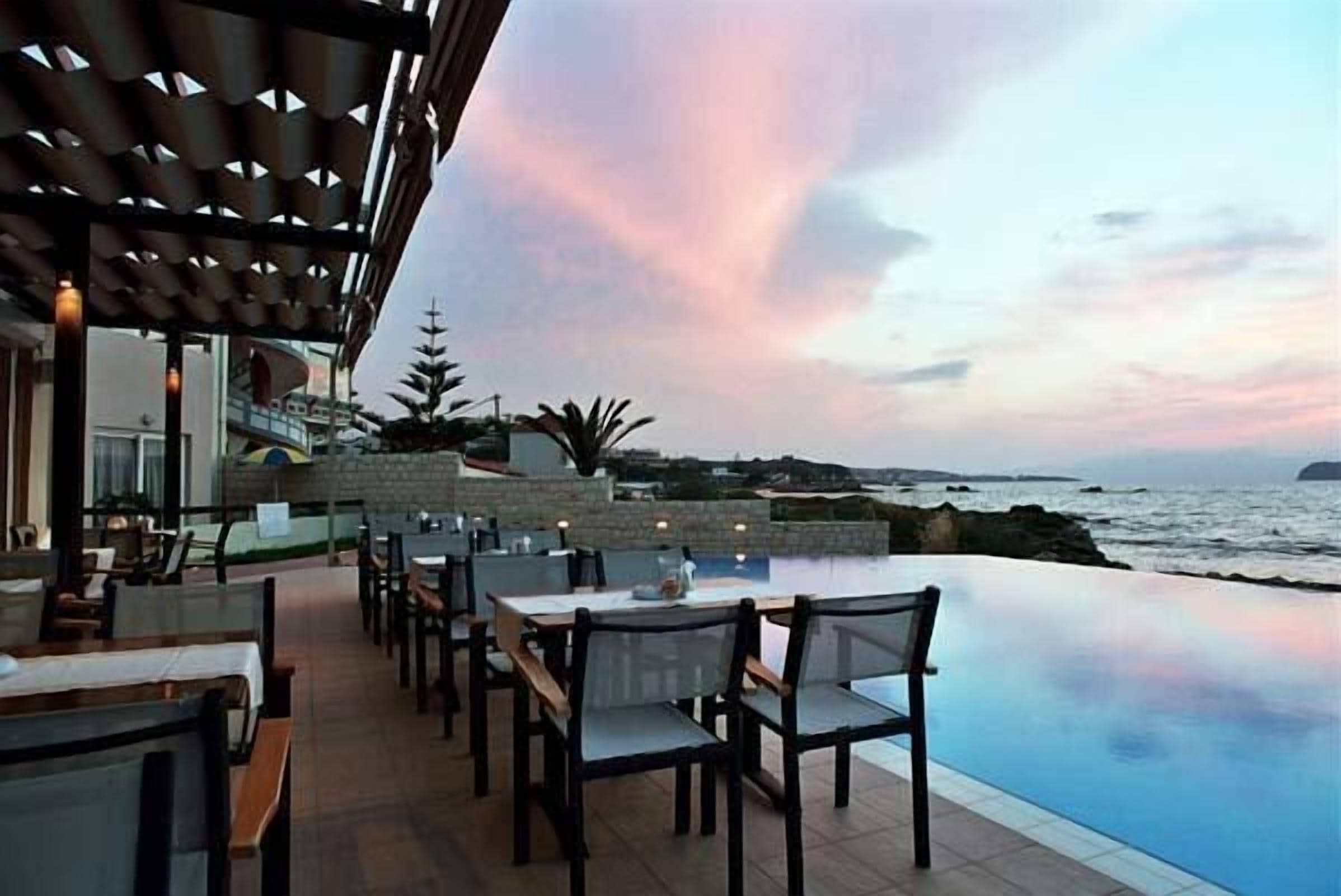 An outdoor dining area with tables and chairs, overlooking a body of water and a sunset sky.
