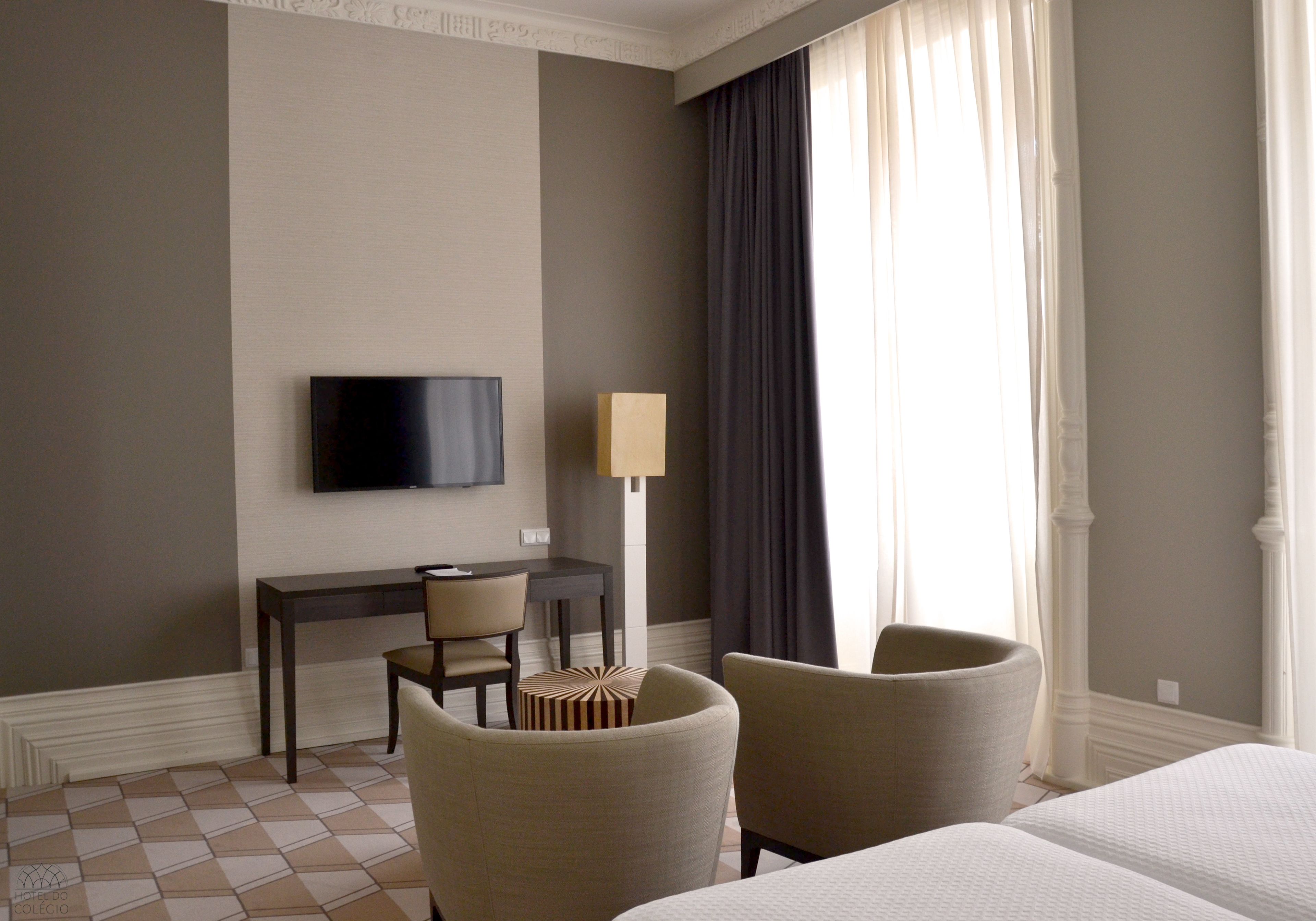 A hotel room with a bed, two armchairs, a desk with a chair, a TV mounted on the wall, and a lamp.