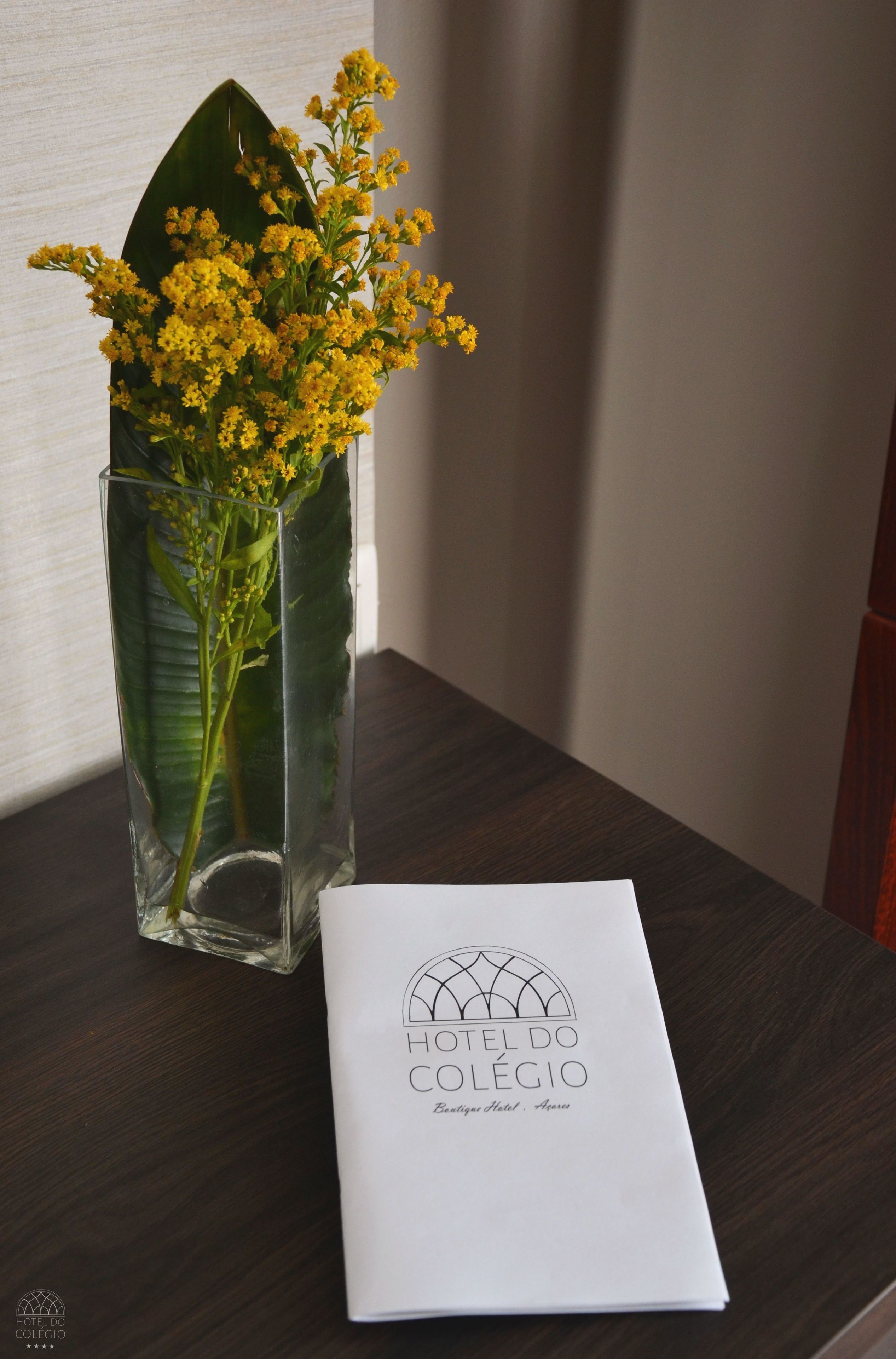 Un jarrón de cristal transparente con flores amarillas y un folleto sobre una mesa de madera.