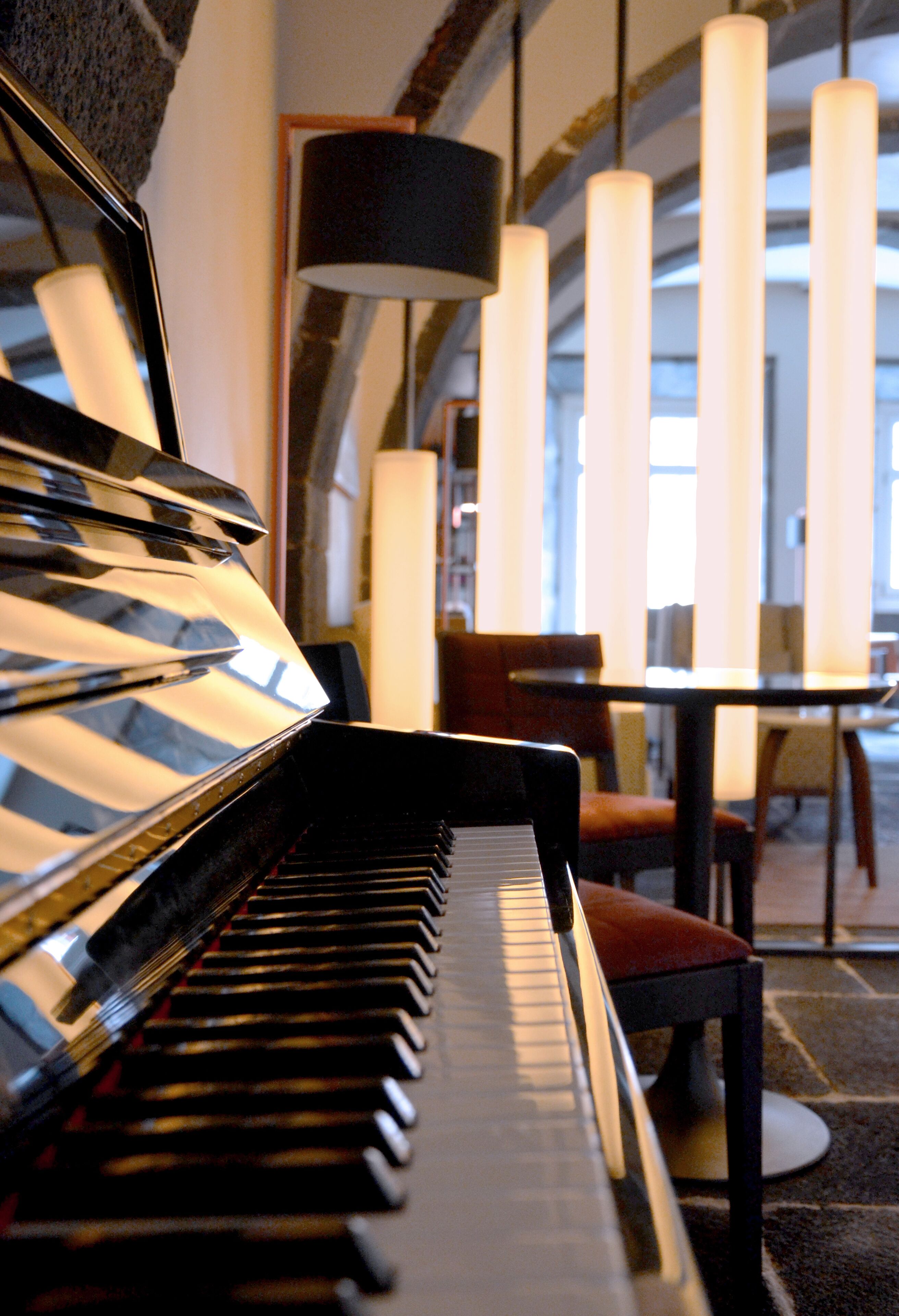 A piano in a room with a table and chairs, large windows, and a lamp.